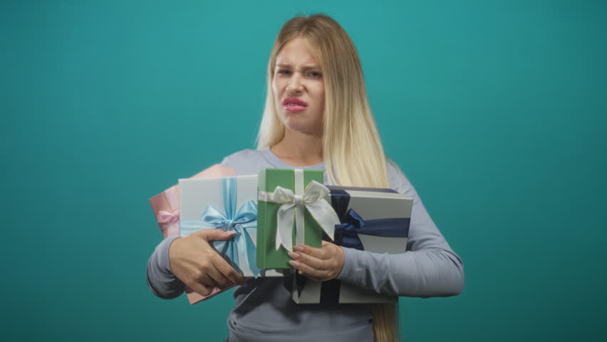 Woman clutching multiple wrapped gift boxes, grimacing with visible face and hands against teal studio backdrop; disgust.