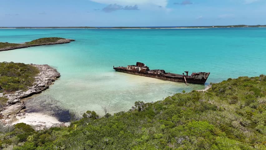 Aerial view of a rusty shipwreck grounded in turquoise waters near the lush green shores of Crab Cay in George Town, Exuma, The Bahamas.