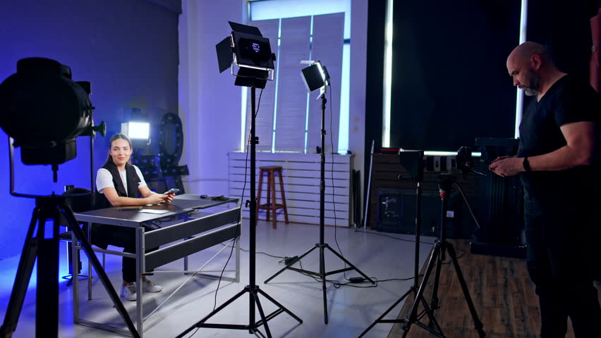 Interior of the modern photo studio with necessary equipment. Smiling lady sits at desk. Cameraman prepares light for footage.