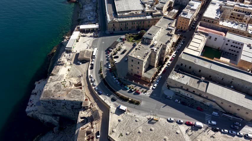 Aerial view of historic stone fortifications and coastal buildings along the waterfront with parked cars on the street in Valletta, Port Region, Malta.