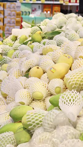 Large pile of fresh mangoes on supermarket fruit display while customer selects ripe tropical fruit in background, grocery shopping scene with produce section and healthy food choice.