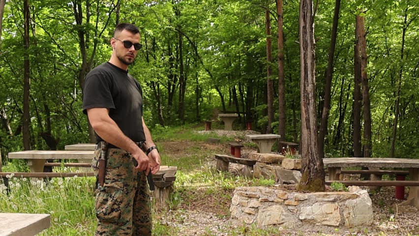 Young man in tactical military style clothing practicing airsoft pistol shooting in a forest recreation area. Focused participant raises and fires an airsoft handgun during outdoor target training.