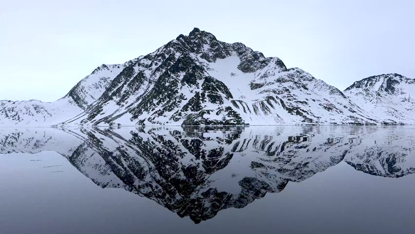 Aerial view of a snow-covered mountain peak perfectly reflected in the calm waters of a fjord in Tasiilaq, Sermersooq Municipality, Greenland.