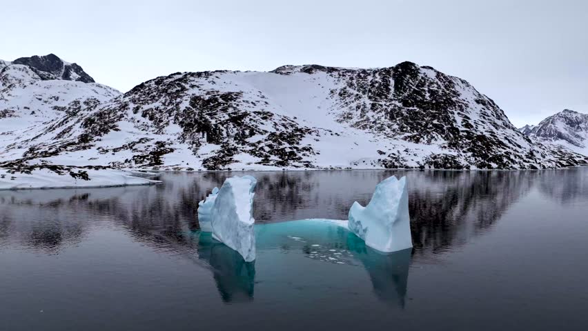 Aerial view of a large iceberg floating in the calm waters of a fjord surrounded by snow-covered mountains in Tasiilaq, Sermersooq Municipality, Greenland.