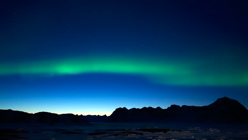 Aerial view of the Aurora Borealis dancing over the silhouette of rugged mountains and ice-covered waters in Tasiilaq, Sermersooq Municipality, Greenland.