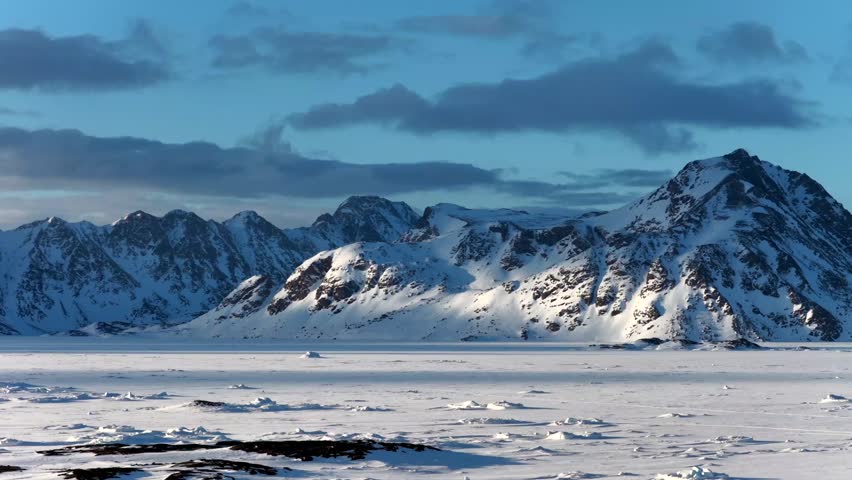Aerial view of snow-covered mountain peaks and a vast frozen ice field under a cloudy blue sky in Tasiilaq, Sermersooq Municipality, Greenland.