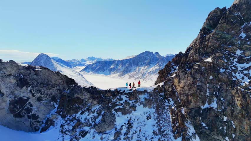 Aerial view of a group of people standing on a snowy mountain ridge overlooking a vast glacier under a clear blue sky in Tasiilaq, Sermersooq Municipality, Greenland.