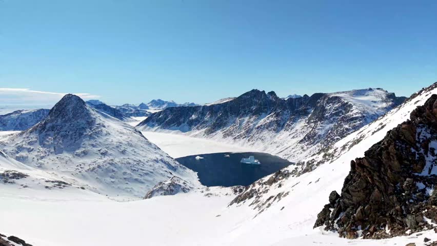 Aerial view of rugged snow-covered mountain peaks and icebergs floating in a dark blue fjord under a clear sky in Tasiilaq, Sermersooq Municipality, Greenland.
