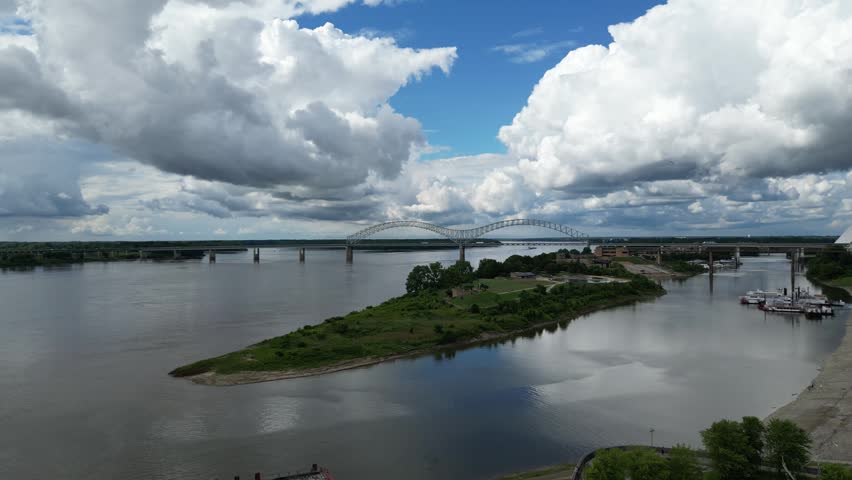 Aerial footage captures a wide river with multiple bridges, a grassy island, and a distinctive pyramid-shaped building on the shore under a cloudy sky.