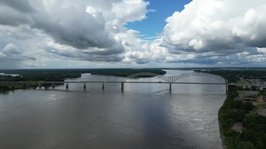 High angle aerial view of a large steel arch bridge spanning a wide river with a boat leaving a white wake in the water below.