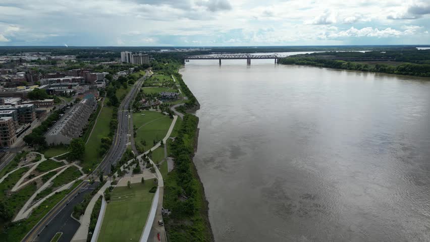 High angle aerial view of a wide river with a truss bridge, adjacent to a green park with winding paths and a city skyline.