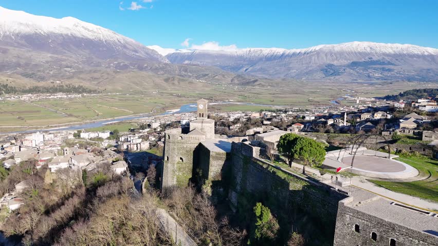 Aerial tracking shot circling the stone walls and clock tower of Gjirokaster Castle, showcasing the valley city and snow-capped mountains of Albania