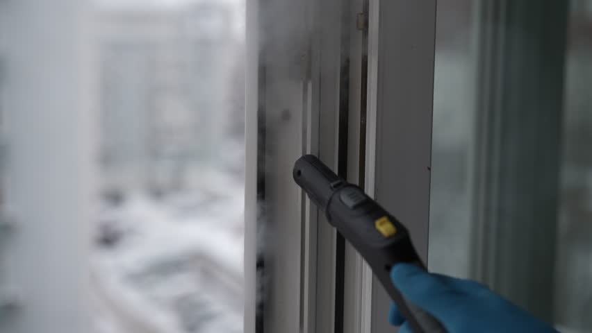 Janitorial service worker deep cleaning dirty window frame with steam generator. Person wearing protective gloves using hot vapor to sanitize surface before wiping it dry. Shooting in slow motion.