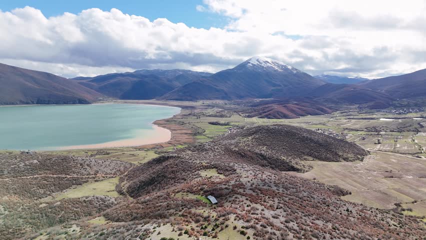 Aerial drone camera moving forward high over Lake Prespa, North Macedonia, revealing green agricultural plains, rural wetlands, and a dramatic snow-covered mountain peak
