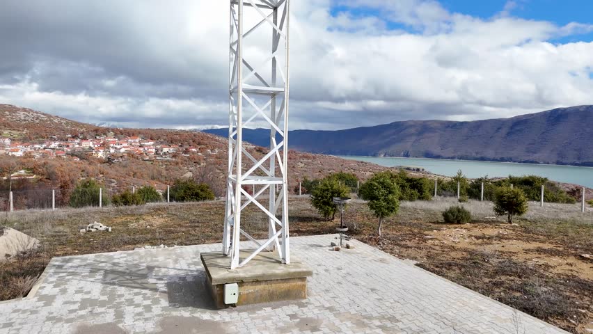 Aerial tracking shot raising upward behind a large white steel lattice cross monument on a hill, overlooking a lakeside village, rugged hills, and the calm waters of Lake Prespa