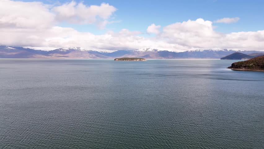 Aerial tracking shot flying forward over the vast waters of Lake Prespa toward the lonely, cliff-bound Maligrad Island with snow-capped mountain ranges behind