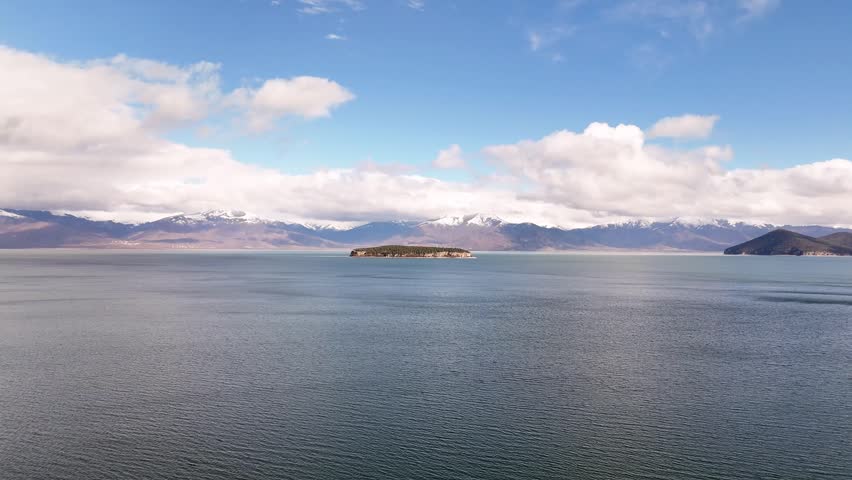 High-angle aerial drone shot circling over the wide waters of Lake Prespa, showcasing the rocky cliffs of remote Maligrad Island against a backdrop of snow-capped Albanian peaks