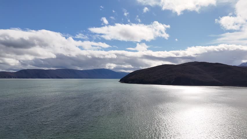 Aerial tracking shot moving forward over the shimmering surface of Lake Prespa, approaching the rocky shores of Maligrad Island under a bright, cloudy sky in Albania