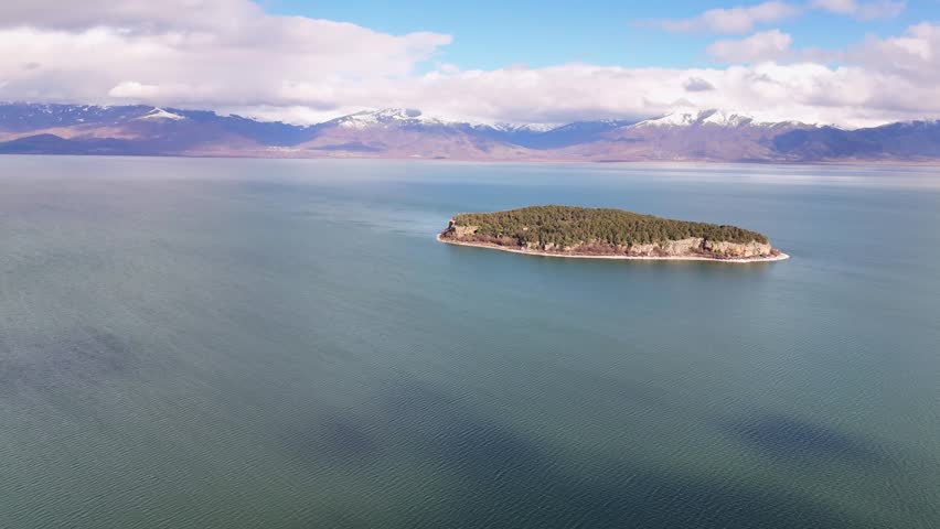 Aerial tracking shot moving right across Lake Prespa, framing the isolated forested shores of Golem Grad Island against a background of snow-capped mountain peaks