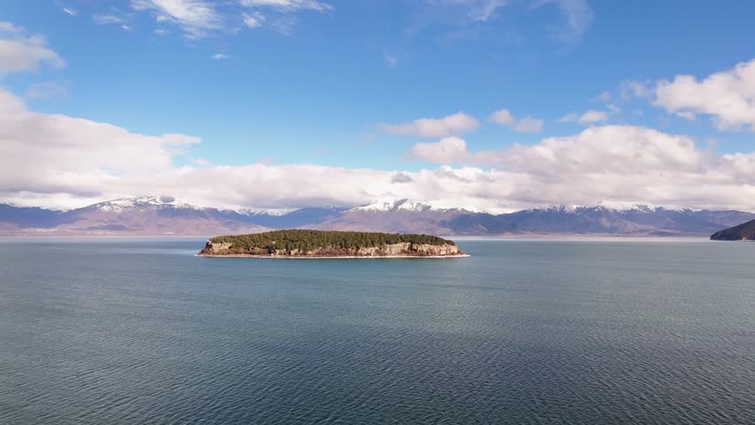 Aerial tracking shot raising upward to reveal the tranquil waters of Lake Prespa, framing the isolated Golem Grad Island and snow-capped mountain ranges under a cloudy blue sky
