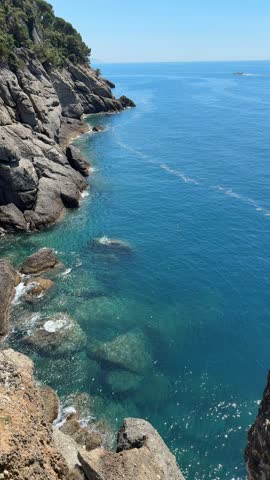 People gather by the rocky coast of Camogli, Italy. The sun shines brightly over the clear blue waters. Gentle waves lap against the stones as boats pass in the distance.