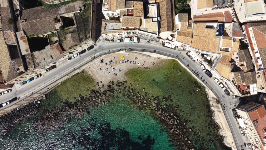 Aerial view of people sunbathing on Cala Rossa Beach surrounded by historic buildings and turquoise water in Syracuse, Sicily, Italy.