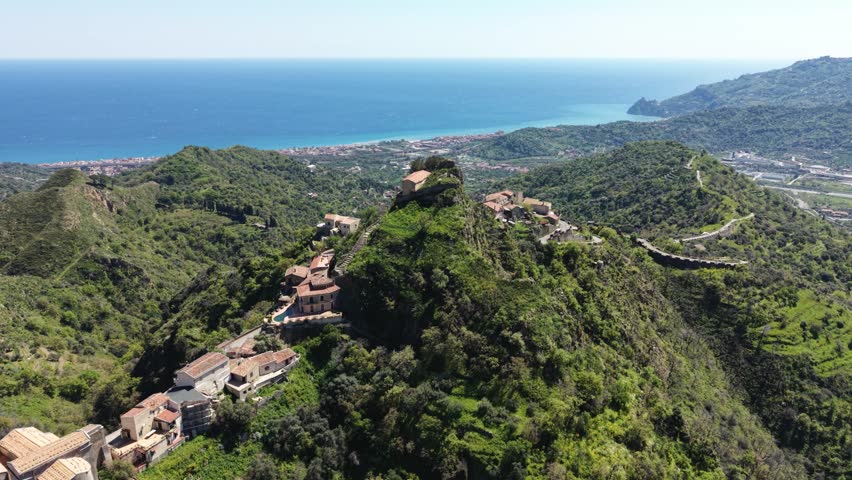 Aerial view of the historic hilltop village of Savoca with stone houses nestled on green ridges overlooking the Mediterranean Sea in Savoca, Sicily, Italy.