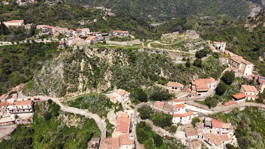 Aerial view of the historic hilltop village of Savoca with stone houses and winding roads nestled among lush green hills and rocky cliffs in Savoca, Sicily, Italy.