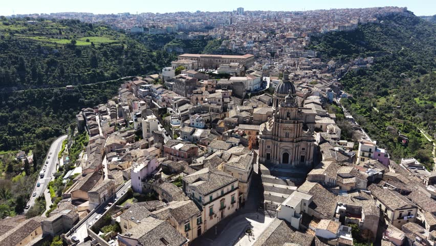 Aerial view of the Cathedral of San Giorgio and the historic baroque architecture of Ragusa Ibla surrounded by lush green hills in Ragusa, Sicily, Italy.