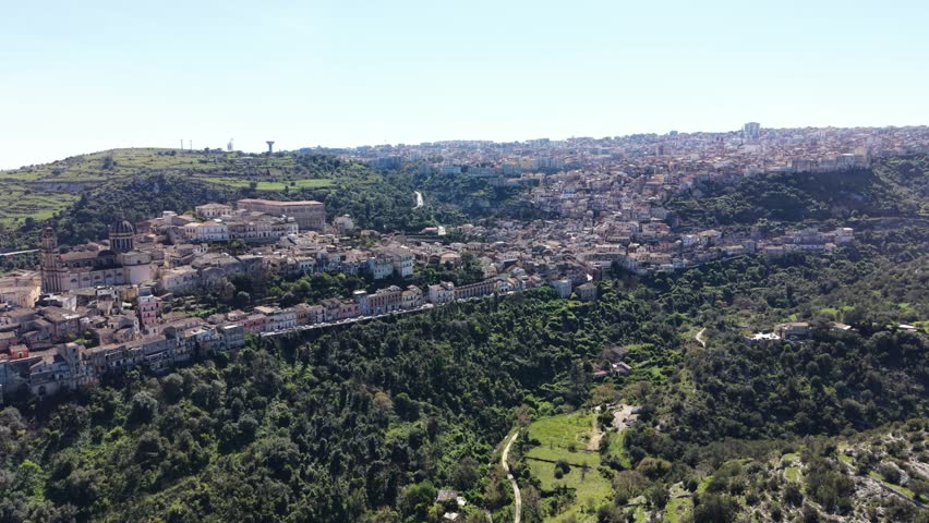 Aerial view of the historic baroque architecture of Ragusa Ibla nestled on a hilltop above a lush green valley under a clear blue sky in Ragusa, Sicily, Italy.
