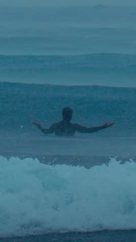 Surfer sitting under stormy rain waiting for wave, dark teal ocean and distant swell, silhouette with arms outstretched, heavy drizzle and foam churn, moody dusk atmosphere, vertical video
