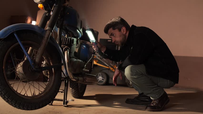 Man crouching and carefully inspecting the engine of a vintage motorcycle using a portable flashlight in a dimly lit home garage, engaged in repair work