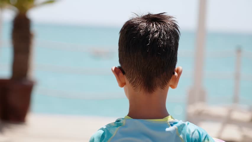 Young child standing barefoot with colorful beach ball on wooden seaside deck, looking at sea during sunny family resort holiday and summer travel. Kid holding ball heads to the edge of pier