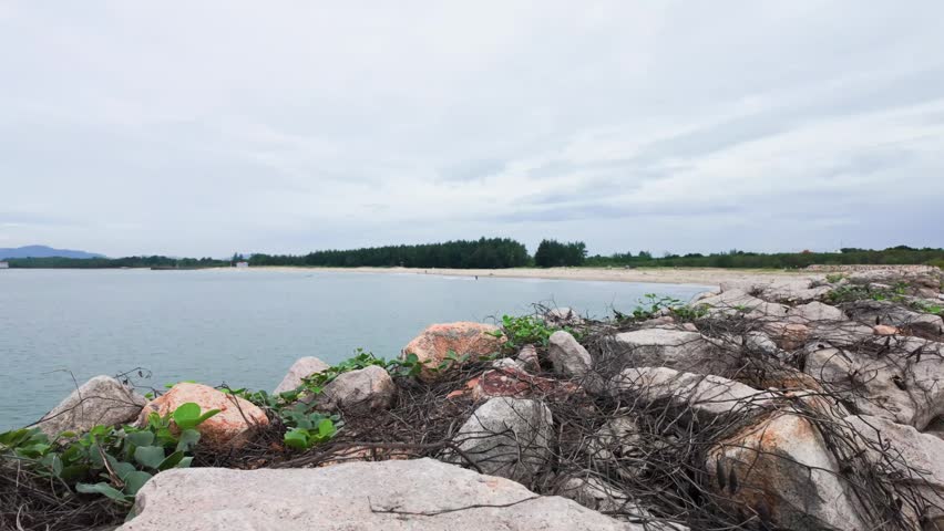 People walk along the riverbank at nuoc ngot pass. The water flows gently beside rocky areas. Trees stand tall in the background under a cloudy sky. The atmosphere feels relaxed and natural.