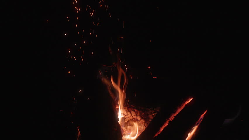 Bright orange sparks rise from a crackling campfire into the pitch black night sky. Burning wood logs glow with intense flames while embers float upward in mesmerizing patterns.