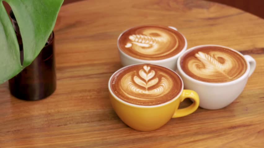 Three cups of warm latte coffee with beautiful and neat latte art on their surfaces. The three cups are placed on a natural brown wooden table, side by side with a vase of green plants on the left side, giving a fresh and aesthetic impression.