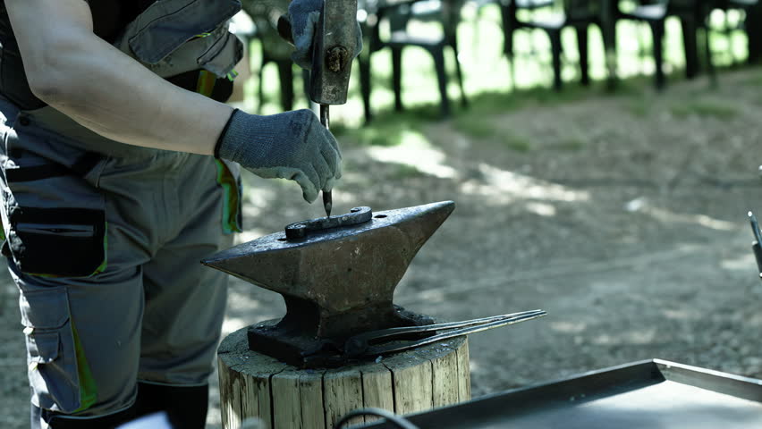 A blacksmith uses a hammer and punch on metal placed on an anvil during outdoor forging work.