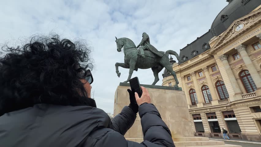 An adult woman uses her smartphone to photograph the Equestrian Statue of Carol I in Bucharest, Romania, against a cloudy sky.