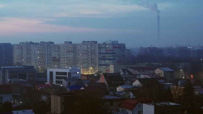 An authentic view of Bucharest at sunrise, featuring residential blocks and houses under a hazy sky. A distant smokestack adds an industrial element to the urban landscape, capturing everyday life.