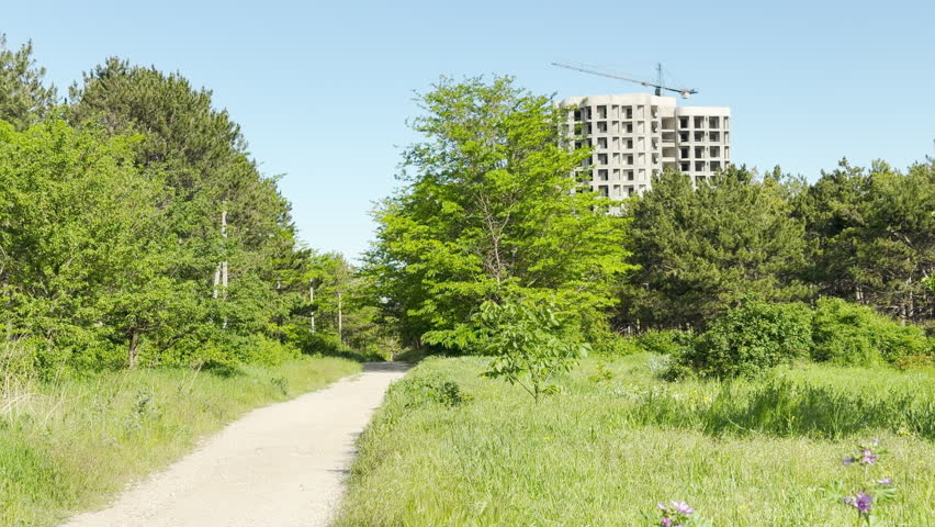 A dirt path leads through a green meadow. An unfinished concrete tower rises above the dense forest. A construction crane stands under a clear sky.