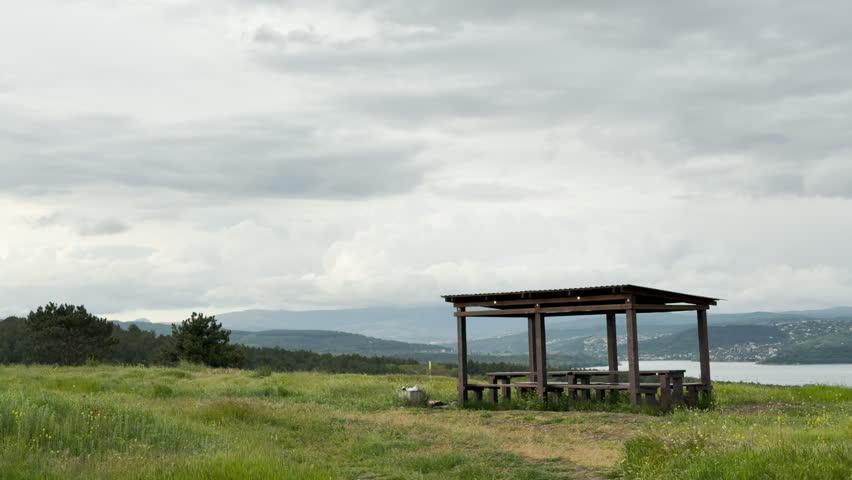 A wooden picnic shelter stands on a grassy hill. The structure overlooks a large lake and rolling mountains under an overcast sky.