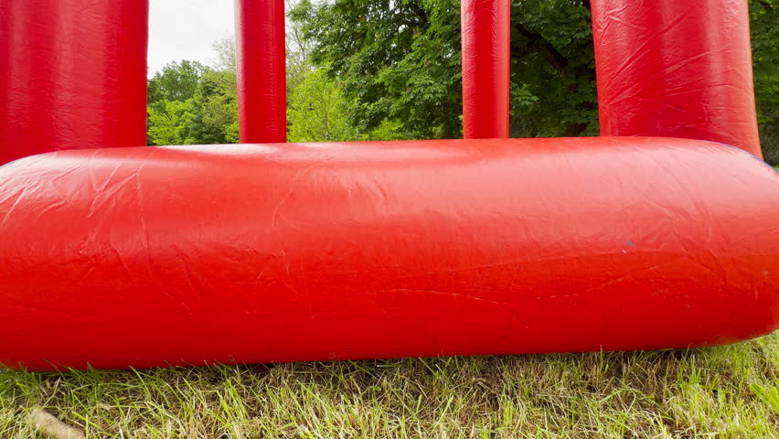 A red and blue inflatable basketball hoop stands tall on the green grass of a park. It is surrounded by lush trees, ready for outdoor fun.
