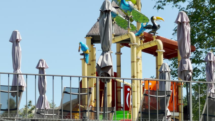 Colorful bird statues sit atop a bamboo-style play structure. Closed patio umbrellas and lounge chairs are arranged on the terrace. The setting is bright and sunny outdoors.