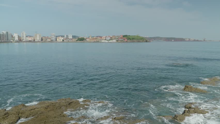 Beautiful wide shot from Playa de San Lorenzo looking across the bay in Gijón, Spain. Features low-tide coastal rocks with white foam and a distant urban city skyline under a clear blue sky.