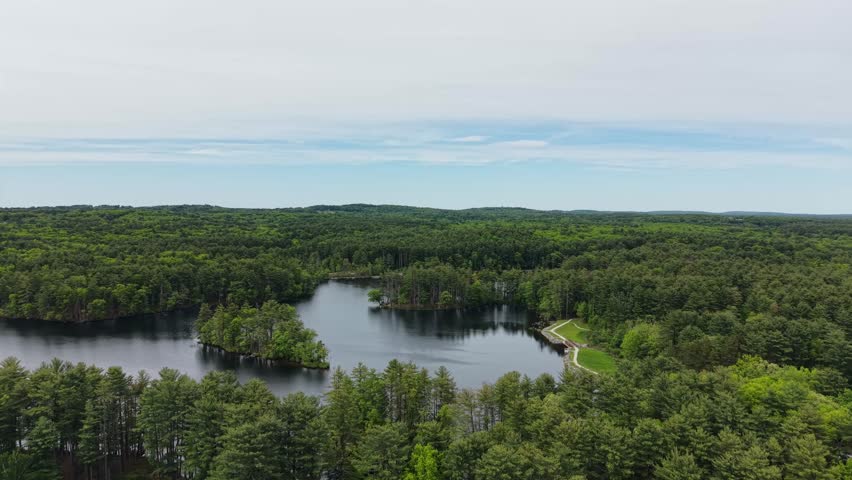 Drone view of dark lake water and small islands surrounded by dense green woodland under cloudy sky in Harold Parker State Forest, Massachusetts, US