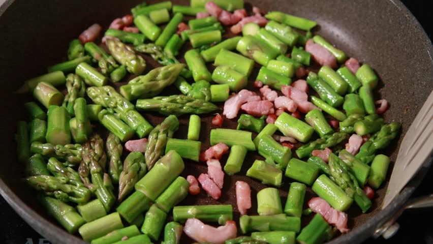 Close-up of fresh green asparagus spears and savory bacon slices sizzling together in a hot frying pan