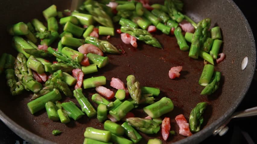 Close-up of fresh green asparagus spears and savory bacon slices sizzling together in a hot frying pan