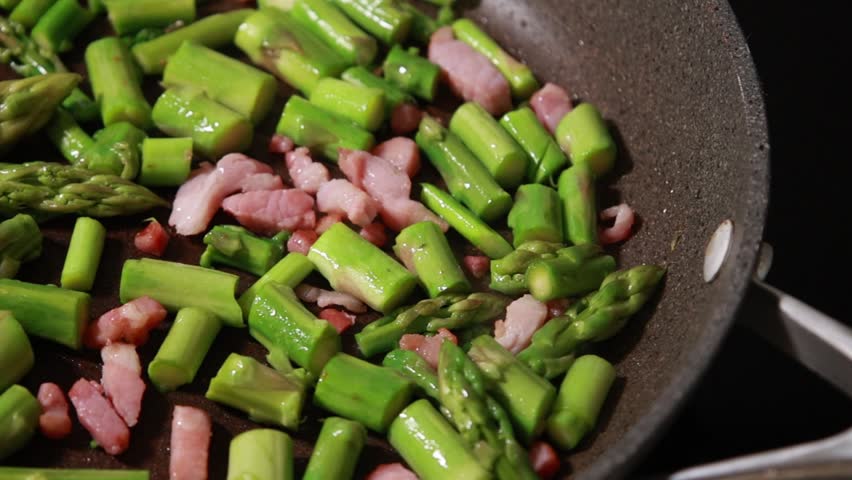 Close-up of fresh green asparagus spears and savory bacon slices sizzling together in a hot frying pan