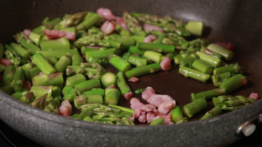 Close-up of fresh green asparagus spears and savory bacon slices sizzling together in a hot frying pan