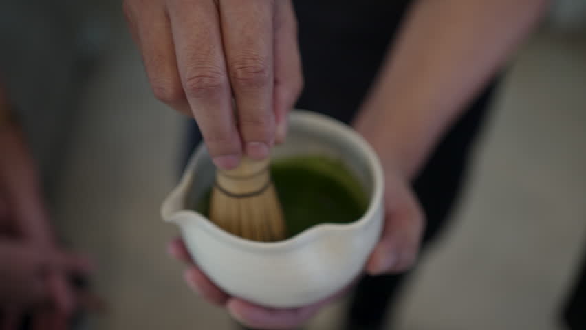 Extreme close-up of matcha green tea being whisked in traditional Japanese tea cup with bamboo tea whisk. Barista preparing matcha at coffee shop.